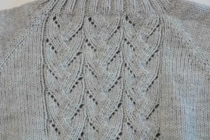 Gros plan réel du motif torsadé ajouré sur le devant du pull gris.